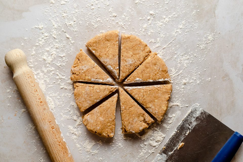 Scone dough rolled into a disc atop a lightly floured creamy marble surface. The dough is cut into 8 wedges. Next to the dough is a rolling pin & a bench scraper.