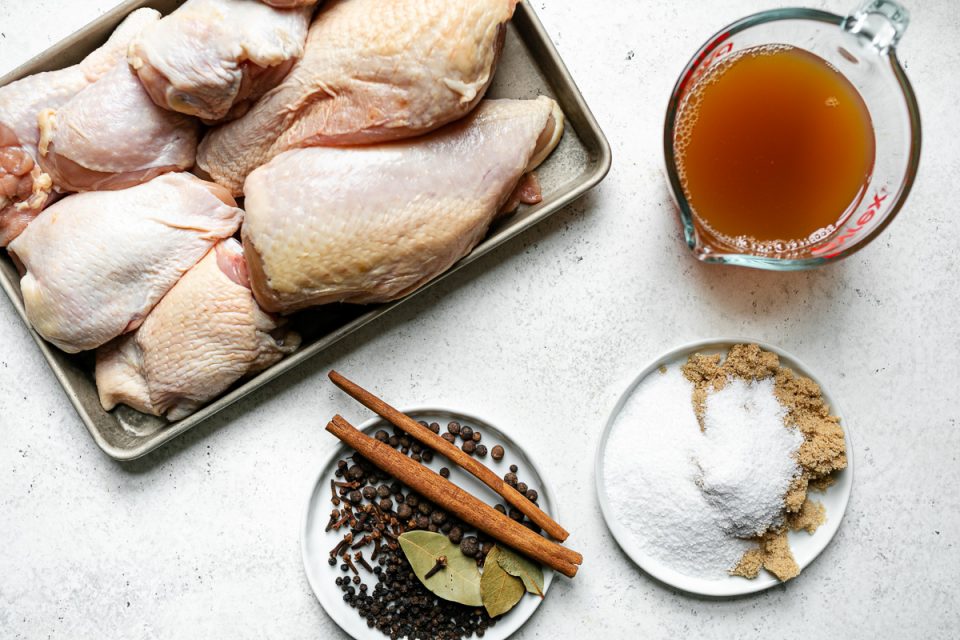 Apple Cider Chicken ingredients arranged on a white surface: a small sheet pan with bone-in skin-on chicken parts, apple cider, salt, brown sugar, whole cinnamon sticks, bay leaves, cloves, & allspice berries.
