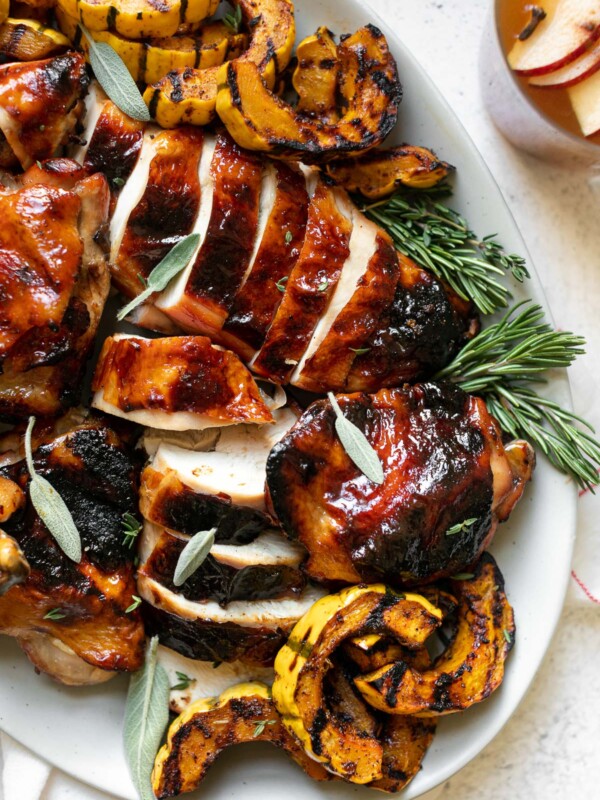 Grilled apple cider chicken on a large serving platter with grilled delicata squash & fresh herbs. The platter sits atop a white surface. Next to the platter is a glass of apple cider (garnished with thinly sliced apple, whole cloves & a cinnamon stick) as well as a red & white striped linen napkin.