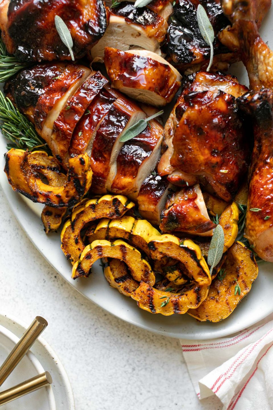 Grilled apple cider chicken on a large serving platter with grilled delicata squash & fresh herbs. The platter sits atop a white surface. Next to the platter is a stack of ceramic plates as well as a red & white striped linen napkin.