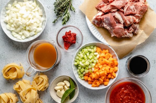 Lamb ragu ingredients arranged on a light blue surface: dried pappardelle nests, onion, beef stock, garlic, bay leaves, tomato paste, crushed tomatoes, celery, carrots, wine, herbs & lamb shoulder.