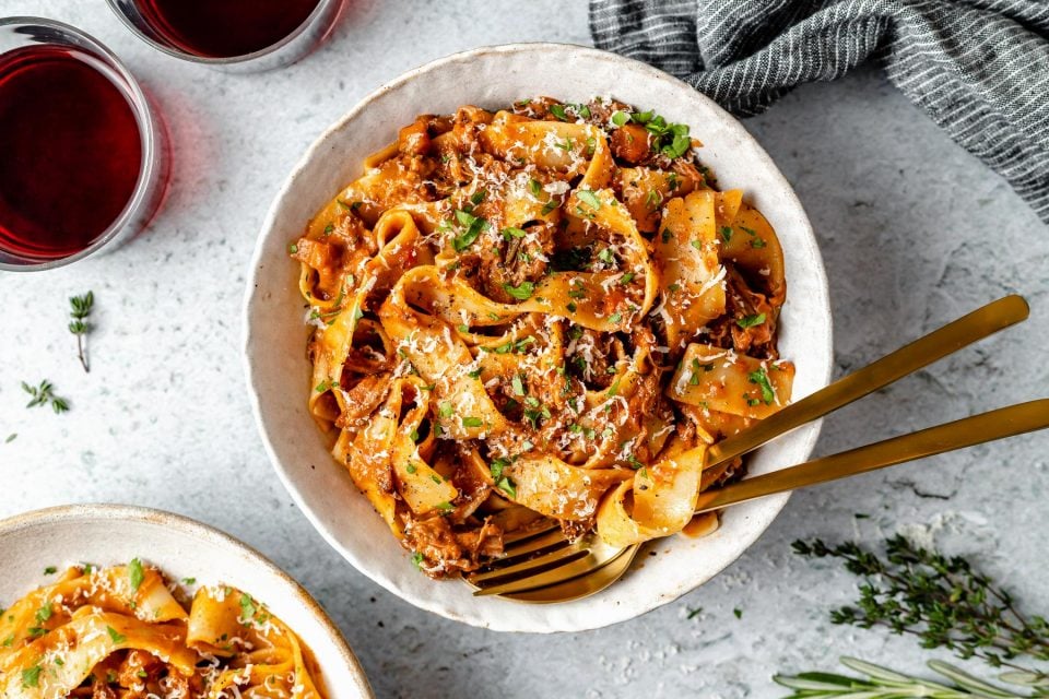 Braised Lamb Ragu Pappardelle shown in pasta bowls, topped with grated parmesan & fresh parsley. The bowls sit atop a light blue surface next to fresh herbs, 2 glasses of red wine & a grey striped linen napkin.