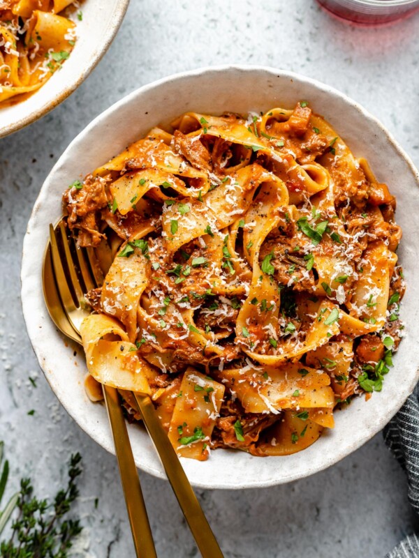 Braised Lamb Ragu Pappardelle shown in pasta bowls, topped with grated parmesan & fresh parsley. The bowls sit atop a light blue surface next to fresh herbs, 2 glasses of red wine & a grey striped linen napkin.