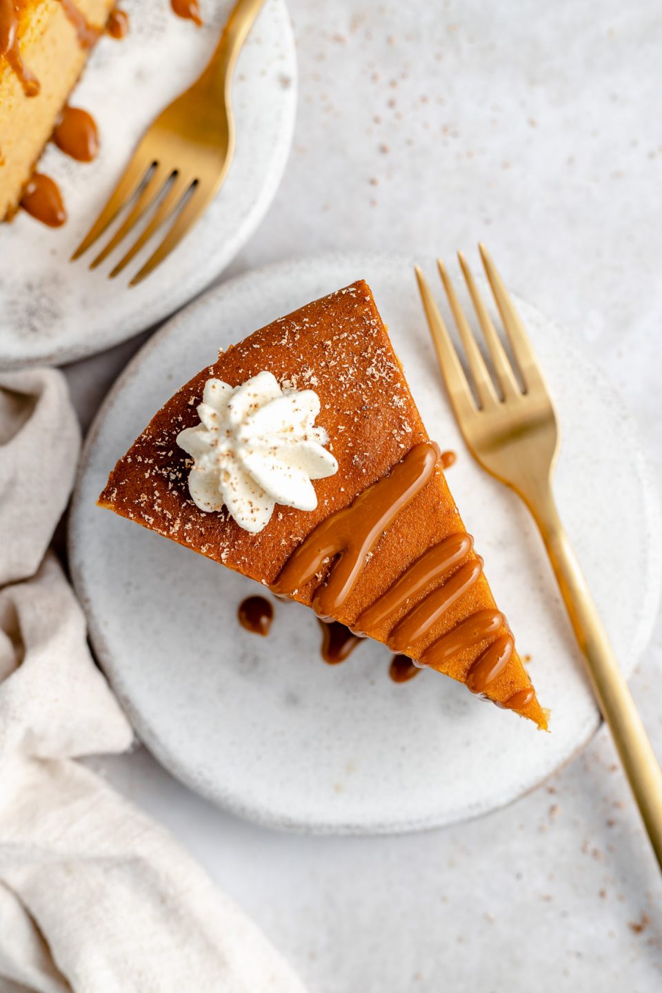 Overhead angle of sliced pumpkin cheesecake, served on a small ceramic plate. The cheesecake is topped with salted caramel, fresh whipped cream, & grated nutmeg. Sitting next to the cheesecake is a large gold fork. A second plate of cheesecake peeks into the top of the frame.