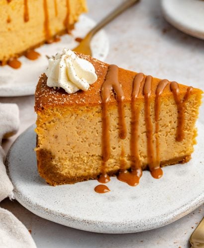 Side angle of pumpkin cheesecake with gingersnap crust. The cheesecake is on a small white ceramic plate, topped with drizzly caramel & freshly whipped cream.