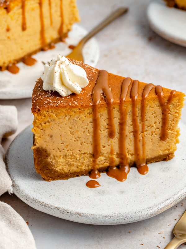 Side angle of pumpkin cheesecake with gingersnap crust. The cheesecake is on a small white ceramic plate, topped with drizzly caramel & freshly whipped cream.