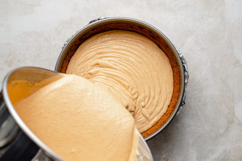 Pouring pumpkin cheesecake filling from large stainless steel mixing bowl into par-baked gingersnap crust, formed in a springform pan.