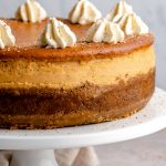 Close up of baked pumpkin cheesecake. The cheesecake is on a white cake stand, topped with piped whipped cream & freshly grated nutmeg.