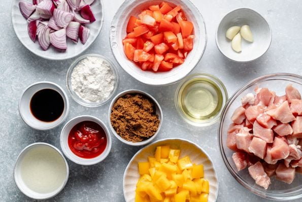 Sweet and sour pork ingredients arranged on a light blue surface in individual bowls & dishes: red onion, rice flour, soy sauce, ketchup, brown sugar, rice vinegar, oil, garlic, tomatoes, bell peppers, & diced pork loin.