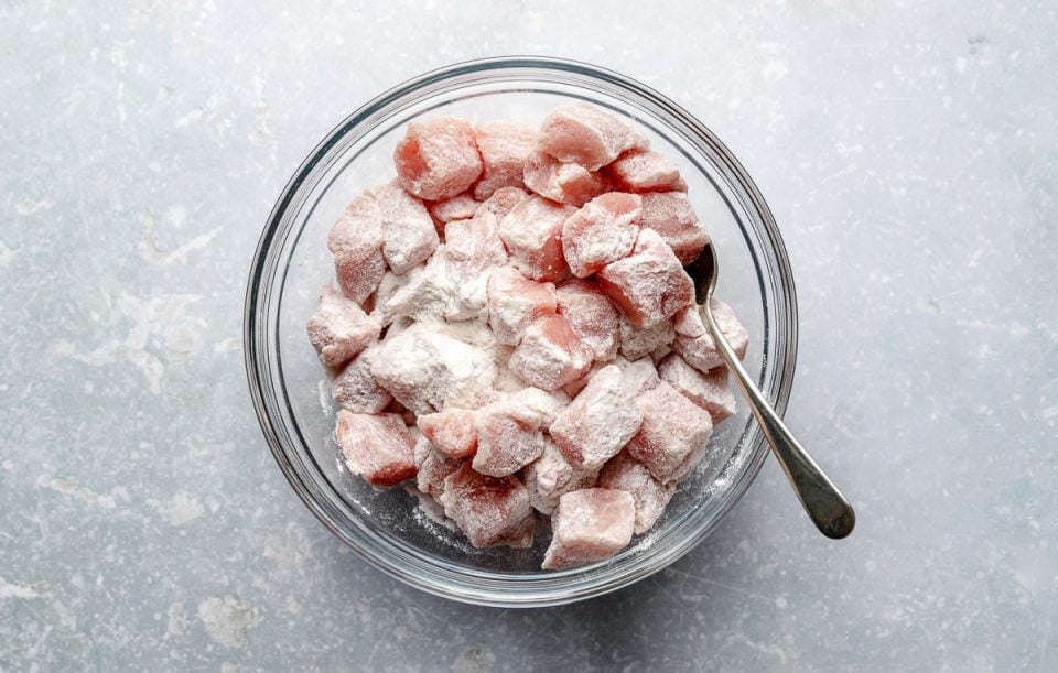 Diced pork loin tossed in rice flour in a large glass mixing bowl atop a light blue surface.