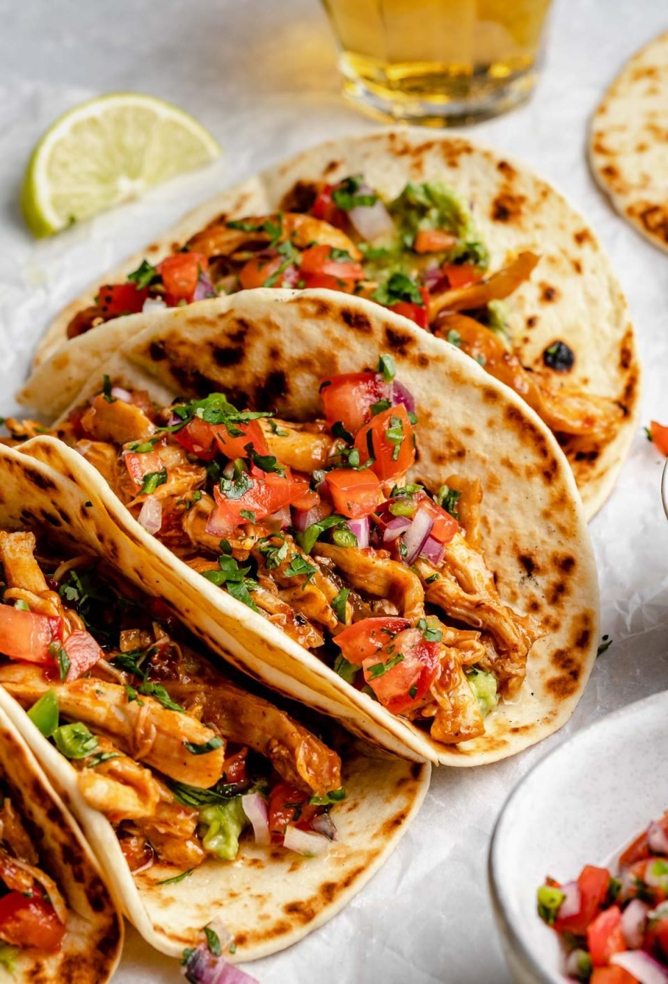 Side angle of chipotle honey chicken tacos pressed together side-by-side on a piece of white crinkled parchment paper. The tacos are garnished with fresh pico de gallo & chopped cilantro. A glass of beer, a lime wedge, & a bowl of pico de gallo surround the tacos.