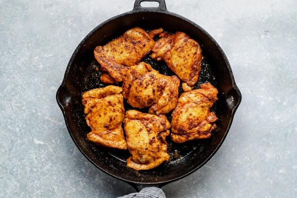 Six chicken thighs seasoned with taco seasoning brown in a large skillet. The skillet rests on a light blue textured surface. The skillet handle has a white and blue linen napkin tied around it.