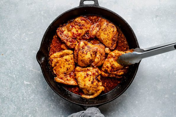 Six pieces of browned chicken thighs rest in a large skillet covered with honey chipotle sauce. The skillet rests on a light blue textured surface. The skillet handle has a white and blue linen napkin tied around it & a pair of tongs is picking up one of the pieces of chicken.