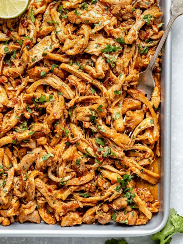 A silver fork scoops up shredded chipotle honey chicken on a small silver baking sheet a top a light blue textured surface. The shredded chicken is garnished with fresh chopped cilantro. A halved lime slice rests on the chicken and a few springs of fresh cilantro rest on the surface in the foreground.