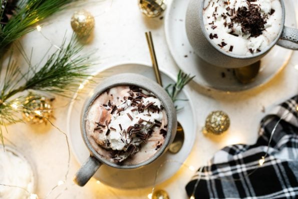 Christmas hot chocolate shown in 2 gray mugs, which sit atop 2 white saucers. The cocoa is topped with freshly whipped cream & shaved chocolate. The saucers sit atop a white surface, next to a black plaid linen napkin, twinkly lights, Christmas ornaments, & decorative pine tree.