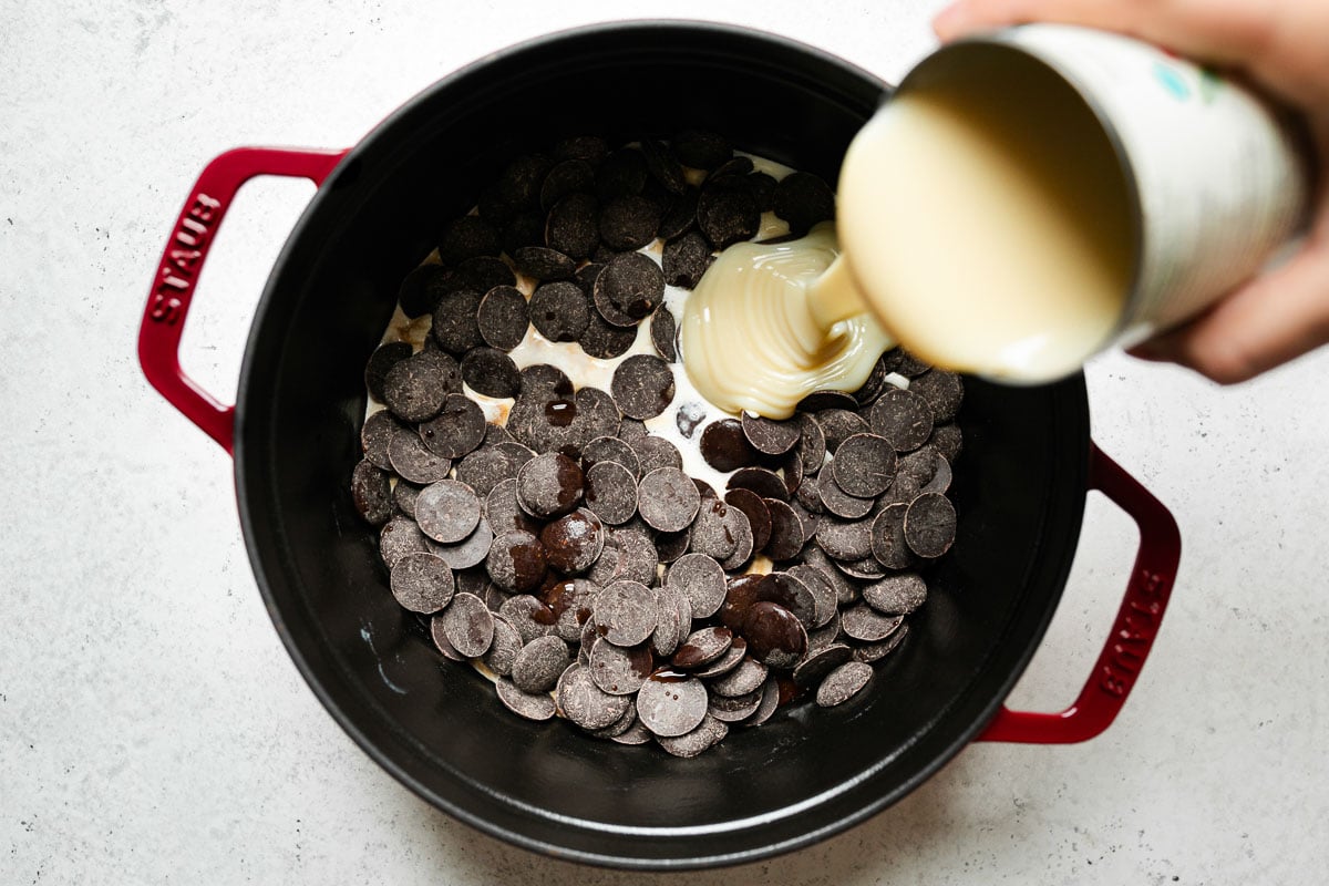 How to make homemade hot cocoa: milk, chocolate wafers & vanilla extract in a red Staub dutch oven. A woman's hand pours a can of sweetened condensed milk into the dutch oven. The dutch oven sits atop a white surface.