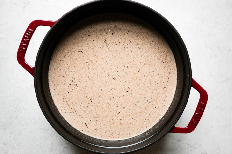 How to make homemade hot cocoa: chocolate partially melted (even more) into the milk mixture in a red Staub dutch oven, which sits atop a white surface.