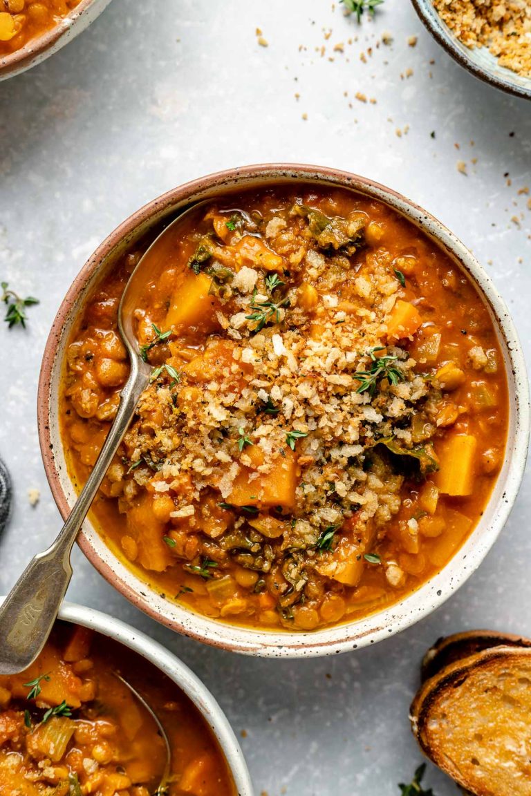 Vegan lentil soup, topped with toasted breadcrumbs, served in 3 speckled ceramic bowls. The bowls sit atop a light blue surface. The bowls are surrounded by crusty buttered bread, spoons, and a sprinkling of fresh herbs & breadcrumbs.