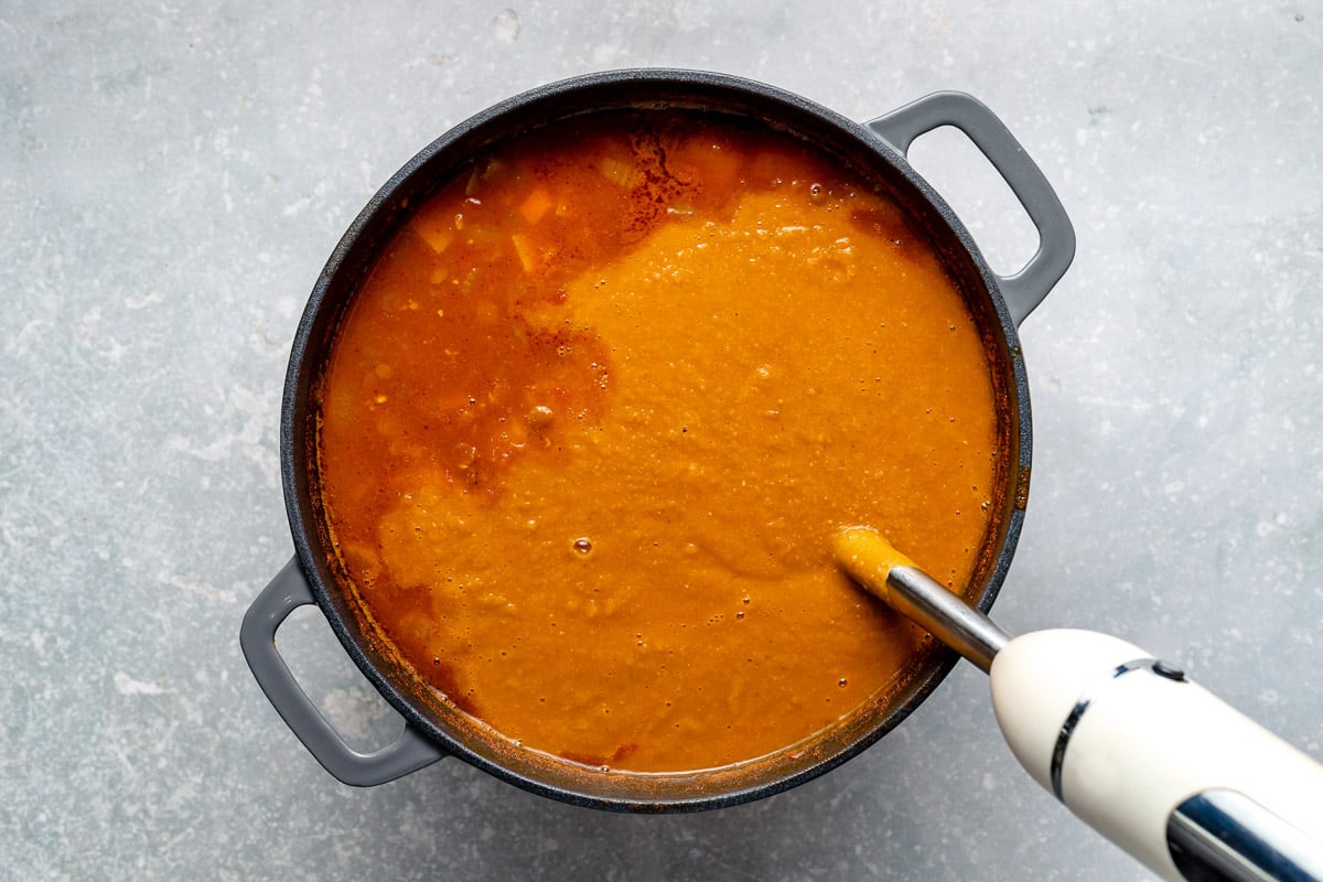 Blending lentil soup: an immersion blender submerged in lentil soup in large grey dutch oven atop a light blue surface.