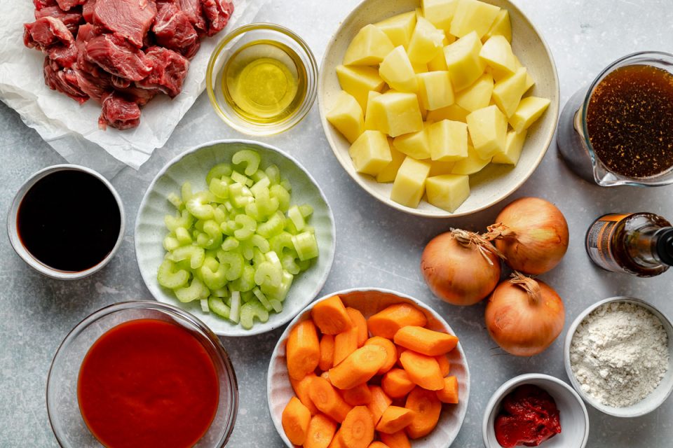 Hawaiian beef stew ingredients arranged on a light blue surface – cubed beef, soy sauce, tomato sauce, celery, carrots, potatoes, onions, tomato paste, flour, beef stock & Worcestershire.