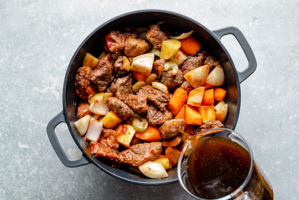 How to make Hawaiian Beef Stew, Step 3: Beef stew ingredients (browned beef, carrots, celery, onion, potatoes) mixed together in a large Dutch oven, which sits atop a light blue surface. Beef stock being poured into the Dutch oven from a large liquid measuring cup.