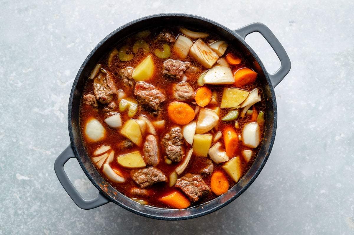 How to make Hawaiian Beef Stew, Step 3: Beef stew ingredients (browned beef, carrots, celery, onion, potatoes, beef stock) mixed together in a large Dutch oven, which sits atop a light blue surface.