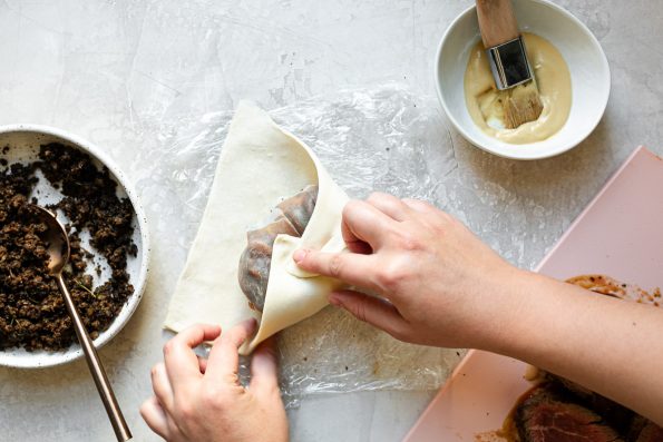 An overhead shot of a woman's hands wrapping puff pastry around prosciutto-wrapped beef tenderloin. A bowl of duxelles, a bowl of dijon mustard and a pink cutting board with beef tenderloin sit on a white surface alongside it.