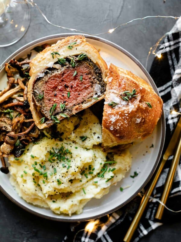 Individual Beef Wellington, cut in half, one half facing up, on a white ceramic plate, served with mashed potatoes and roasted mushrooms. The plate is on a black surface, next to a black plaid linen napkin, gold flatware, a glass of wine and twinkly lights.
