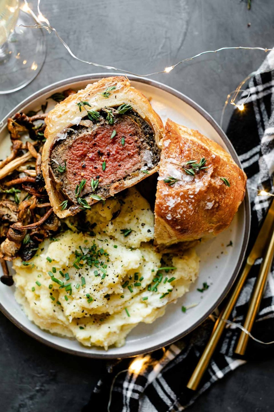 Individual Beef Wellington, cut in half, one half facing up, on a white ceramic plate, served with mashed potatoes and roasted mushrooms. The plate is on a black surface, next to a black plaid linen napkin, gold flatware, a glass of wine and twinkly lights.
