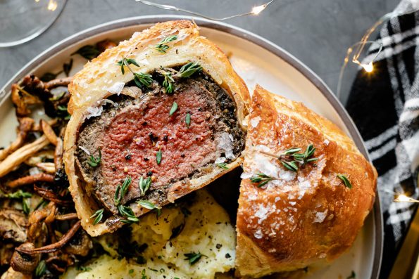 Individual Beef Wellington, cut in half, one half facing up. The beef wellington is on a white ceramic plate, served with mashed potatoes & roasted mushrooms. The plate is on a black surface, next to a black plaid linen napkin, gold flatware, a glass of wine & twinkly lights.