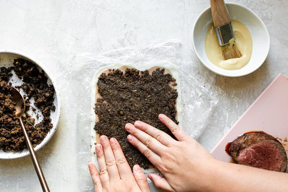 An overhead shot of a woman's hands spreading mushroom duxelles over thinly sliced prosciutto on a white surface. A bowl of duxelles, a bowl of dijon mustard and a pink cutting board with beef tenderloin sit alongside it.