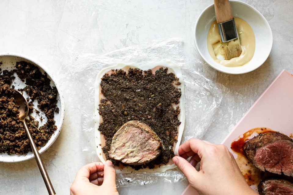 An overhead shot of a woman's hands wrapping Dijon-brushed filet mignon in prosciutto and mushroom duxelles. A bowl of duxelles, a bowl of dijon mustard and a pink cutting board with beef tenderloin sit on a white surface alongside it.