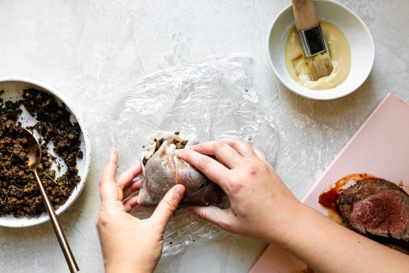 An overhead shot of a woman's hands wrapping Dijon-brushed filet mignon in prosciutto + mushroom duxelles. A bowl of duxelles, a bowl of dijon mustard and a pink cutting board with beef tenderloin sit on a white surface alongside it.