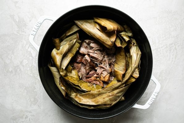 Shredded Kalua pig shown inside dried banana leaf in a white Dutch oven atop a creamy cement surface.