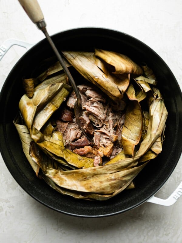 Shredded Kalua pig shown inside dried banana leaf in a white Dutch oven atop a creamy cement surface. A serving fork with a long wooden handle is inserted in the center of the pork.