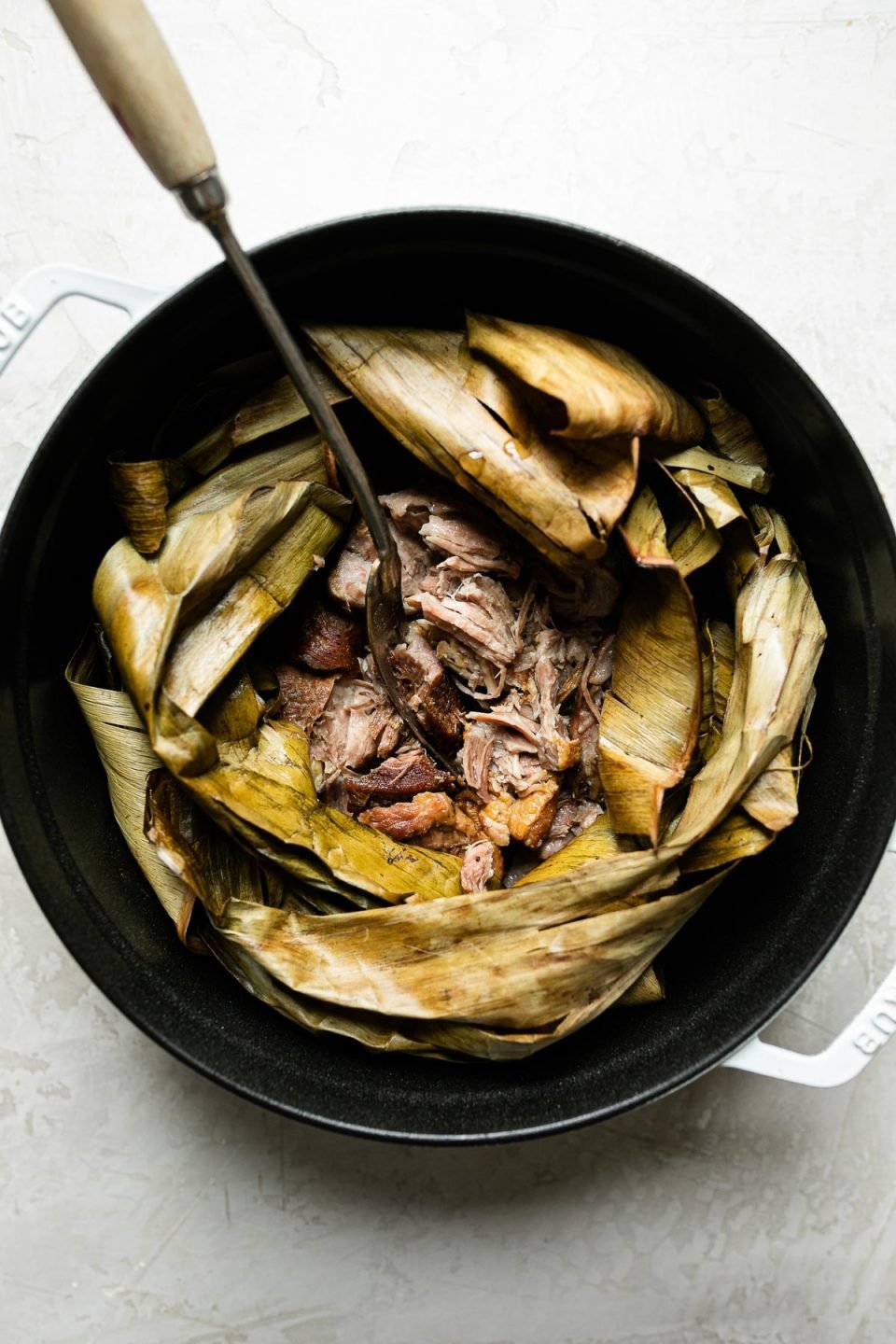 Shredded Kalua pig shown inside dried banana leaf in a white Dutch oven atop a creamy cement surface. A serving fork with a long wooden handle is inserted in the center of the pork.