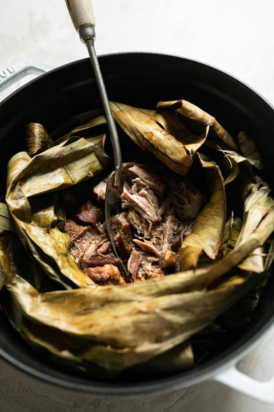 Side angle of shredded Kalua pig shown inside dried banana leaf in a white Dutch oven atop a creamy cement surface. A serving fork with a long wooden handle is inserted in the center of the pork.