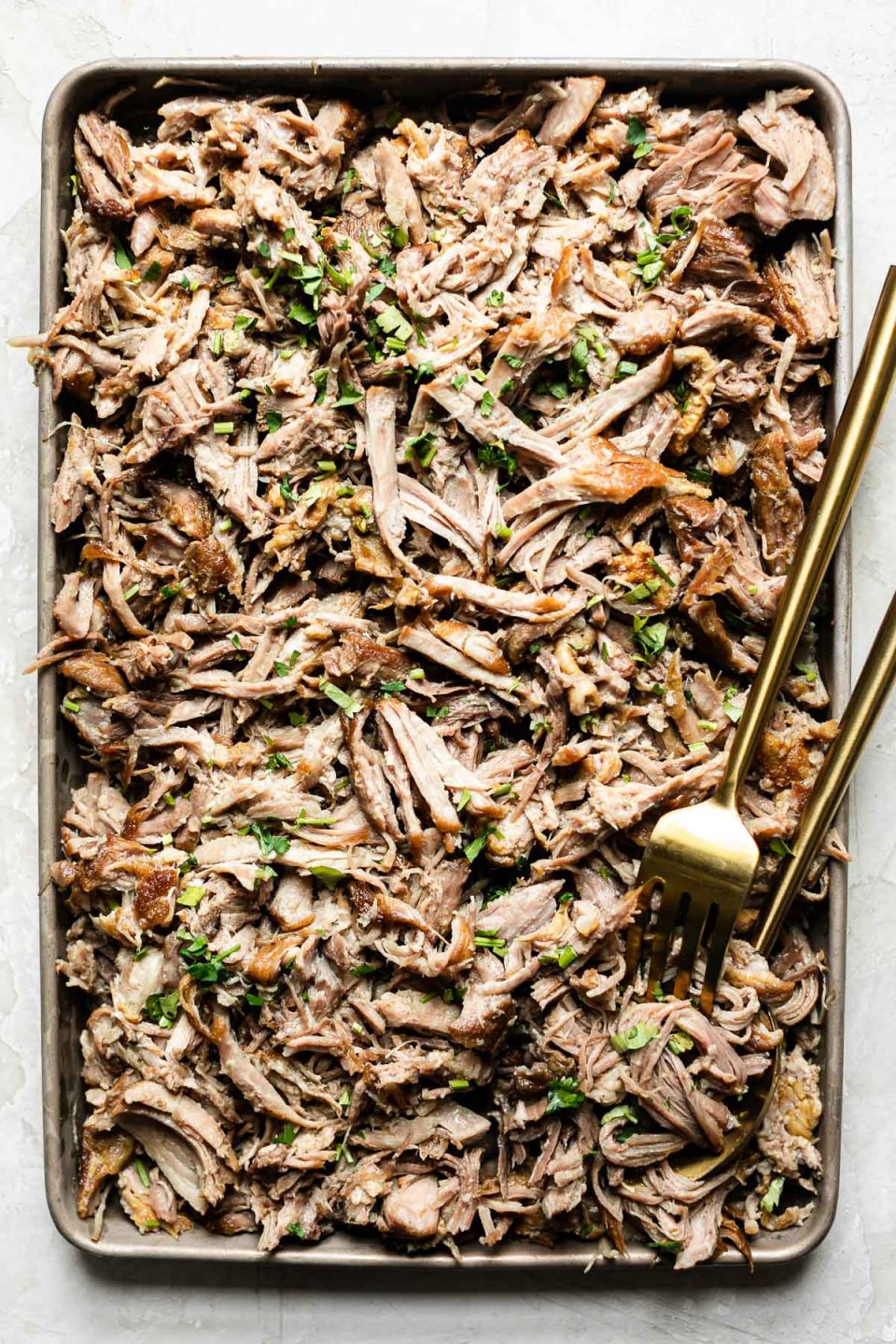 Shredded kalua pork on a small baking sheet, topped with cilantro. A gold serving fork & spoon are nestled in the pork.