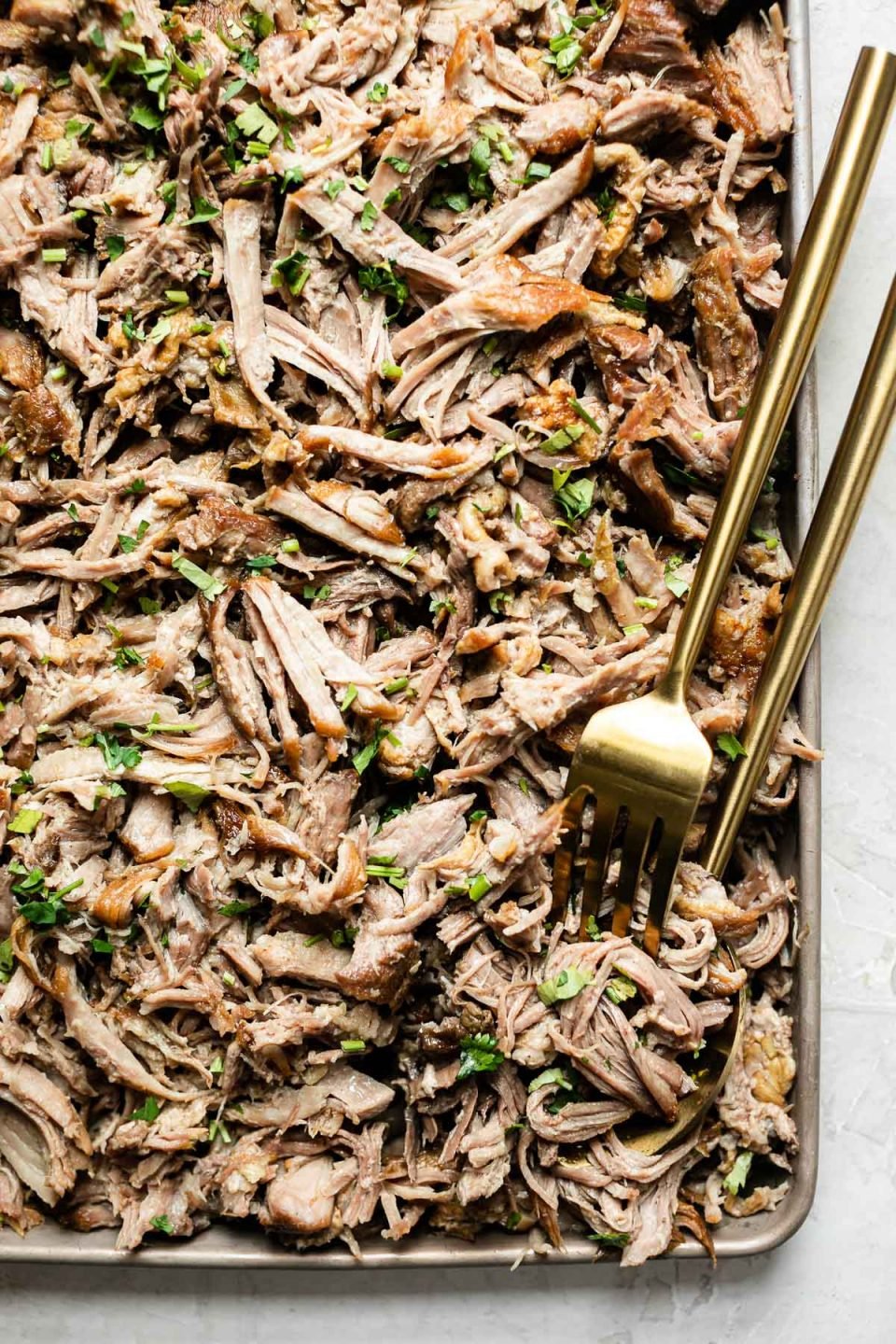 Close up of shredded kalua pork on a small baking sheet, topped with cilantro. A gold serving fork & spoon are nestled in the pork.