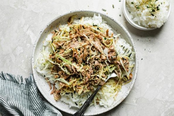 Kalua pork and cabbage served over white rice in a speckled ceramic bowl. The bowl sits atop a cream backdrop, next to a small bowl of white rice & a striped linen napkin.