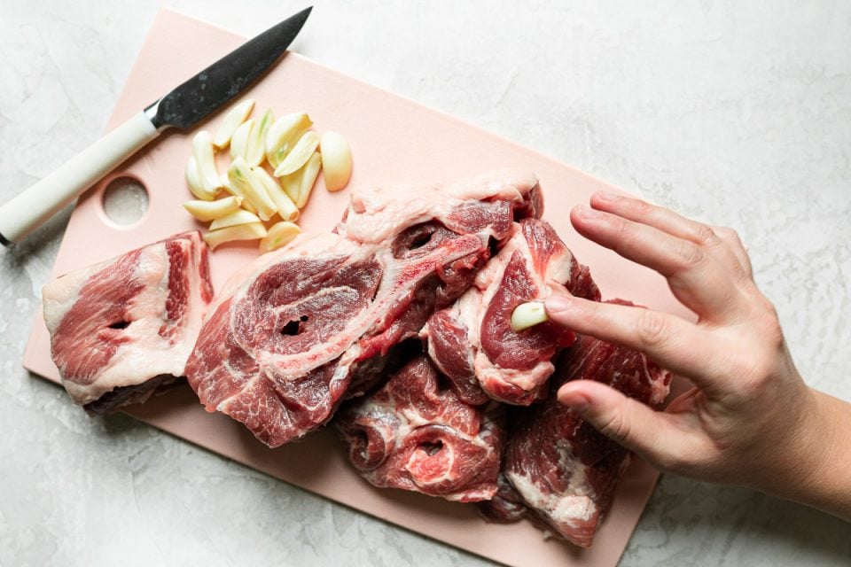 A few large pieces of bone-in pork shoulder sit atop a pink cutting board on a creamy white surface. There are slits cut into the pork & a woman's hand reaches into the frame, pressing garlic into one of the cavities.