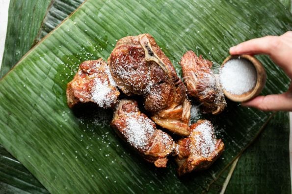 Browned pork shoulder sits atop vibrantly green banana leaf. A woman's hand reaches into the frame with a small bowl of Hawaiian salt, sprinkling the salt over the pork.