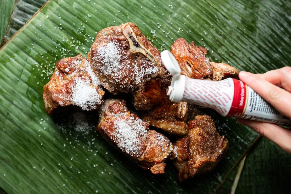 Browned pork shoulder sits atop vibrantly green banana leaf. A woman's hand reaches into the frame with a bottle of Colgin liquid smoke, pouring the liquid smoke over the seasoned pork.