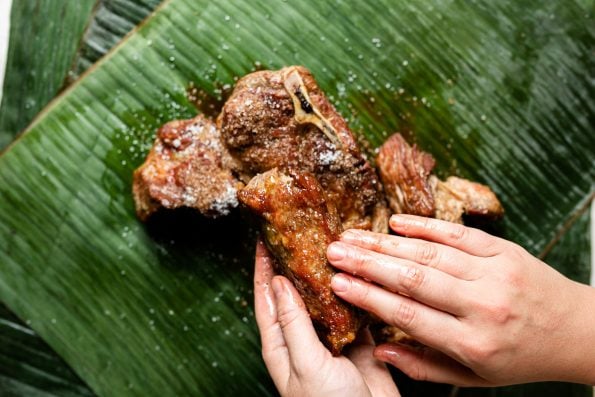 Browned pork shoulder sits atop vibrantly green banana leaf. A woman's hands hold one of the pieces of pork, rubbing the seasoning into the surface.