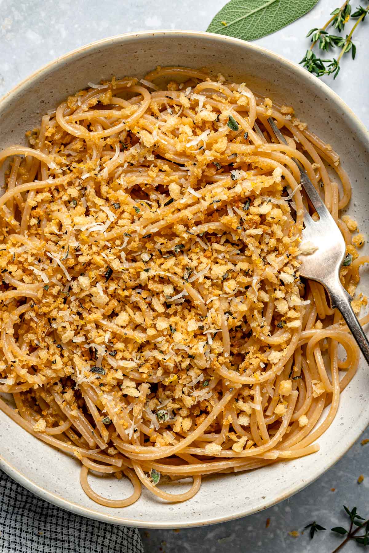 A bowl of spaghetti topped with golden toasted breadcrumbs, grated cheese, and fresh herbs, with a fork resting in the noodles.