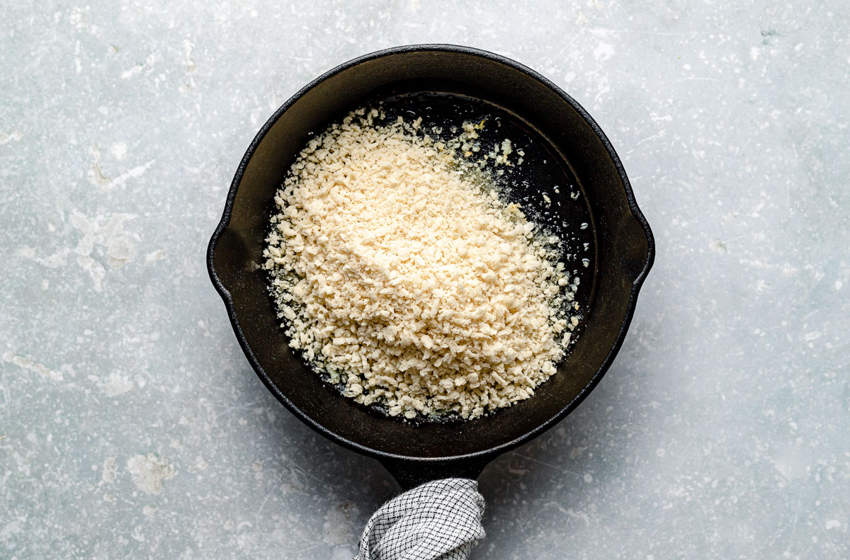 A small black skillet filled with panko breadcrumbs sits on a light gray countertop; the skillet handle is wrapped with a checkered cloth.