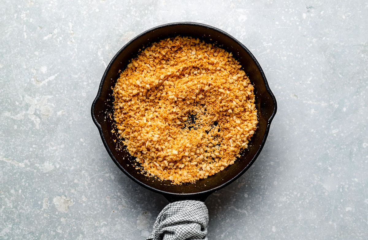 A black skillet filled with golden brown toasted breadcrumbs sits on a light gray textured surface, with a gray cloth wrapped around the skillet handle.