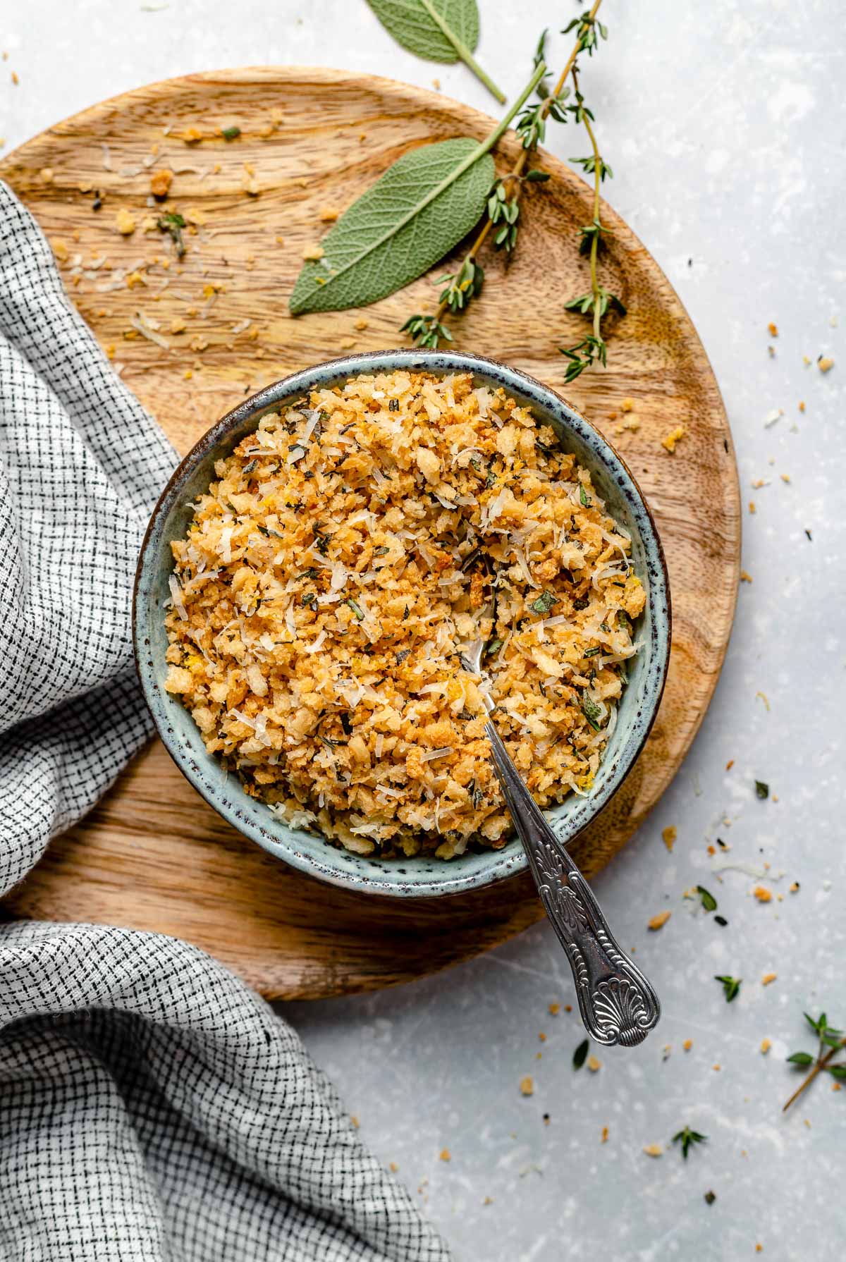 A bowl filled with golden, crumbly stuffing sits on a wooden board, garnished with fresh herbs. A silver spoon rests inside the bowl, and a checkered cloth is draped nearby.