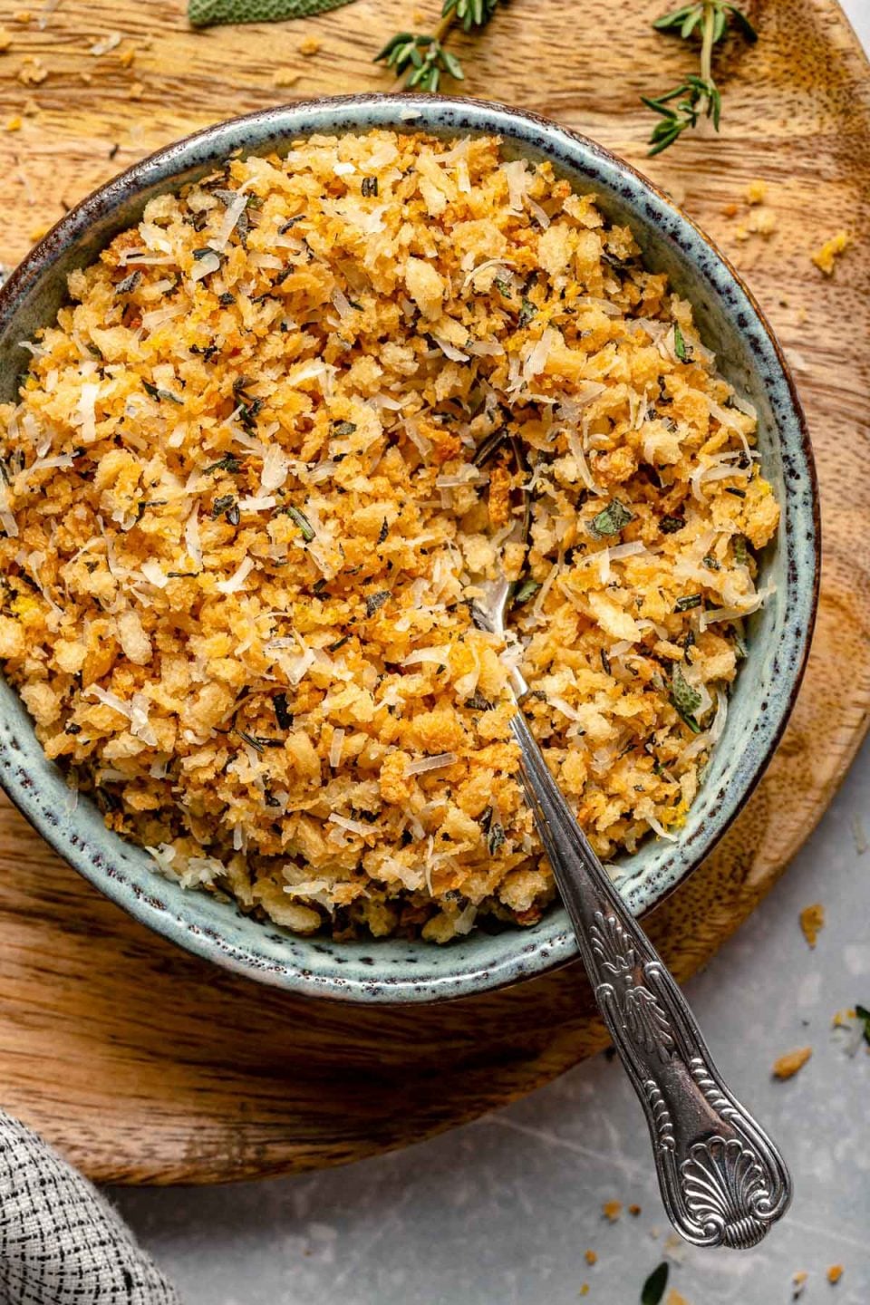 A bowl filled with golden, crispy stuffing topped with herbs, with a spoon resting inside. The bowl is placed on a wooden board, and a few herb sprigs are visible nearby.