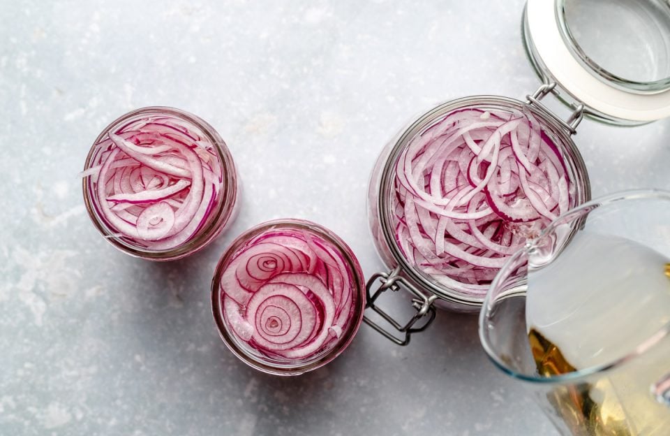 An overhead shot of 3 jars or easy pickled red onions on a light blue surface. Two of the jars are smaller and have been filled with pickling liquid while the third larger jar is getting the pickling liquid poured into it via a liquid measuring cup.